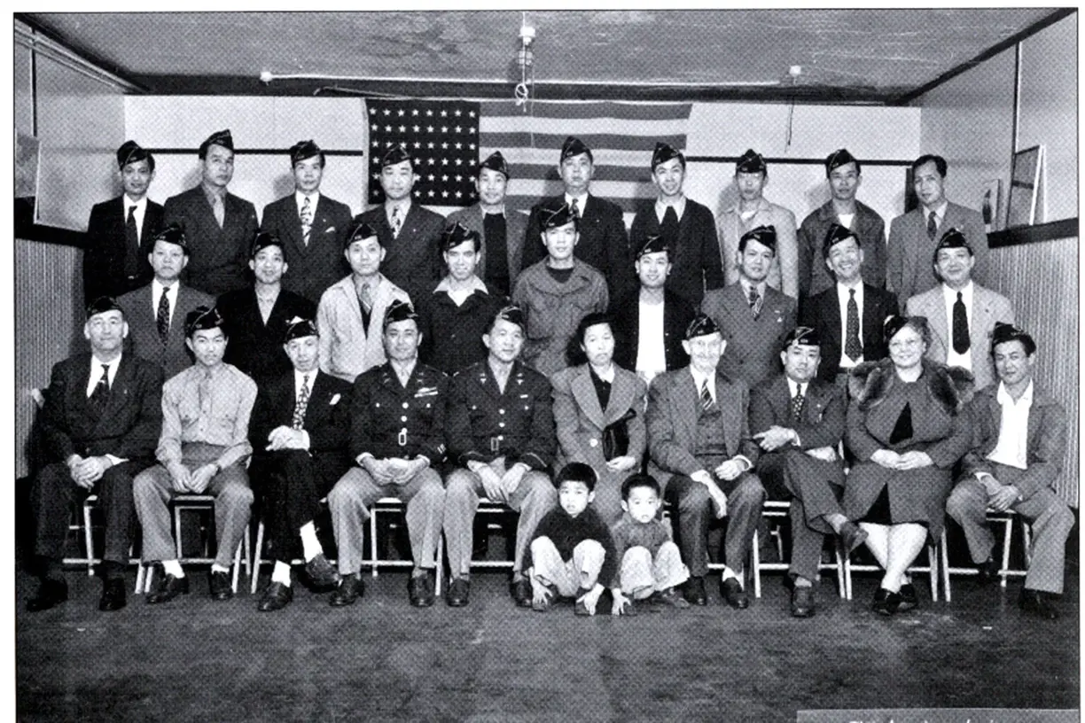 An early group photograph of Chinatown Post 628 veterans and their families, gathered in front of an American flag.